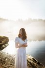 Жінка стоїть в білій сукні на скелі, дивлячись на озеро в туманний день — стокове фото