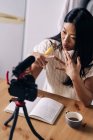 Зверху молода етнічна жінка відеоблогер з блокнотом, що сидить за столом з фотоапаратом на тринозі на кухні — стокове фото