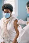 Специалистка-женщина в защитной форме, латексных перчатках и маске для лица, вакцинирующая взрослую афроамериканку во время вспышки коронавируса — стоковое фото