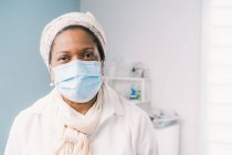 Femme mûre afro-américaine patiente avec masque facial assise au lit de la clinique en attente de rendez-vous pendant l'épidémie de coronavirus — Photo de stock