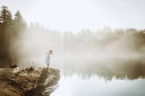 Vue de dos femme debout vêtue d'une robe blanche sur un rocher regardant un lac par une journée brumeuse — Photo de stock