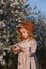 Entzückendes kleines Mädchen in Kleid und Hut, das neben einem Baum mit blühenden Blumen steht und in den Frühlingsgarten schaut — Stockfoto