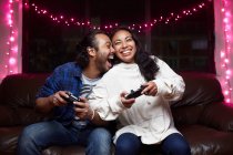 Возбужденная этническая пара в повседневной одежде с джойстиками, играющая в видеоигры вместе, сидя дома на кожаном диване — стоковое фото