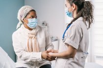 Молодая женщина-врач в медицинской форме и стетоскопе в маске для лица говорит и держит за руки взрослую афро-американскую пациентку во время приема в клинике — стоковое фото