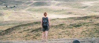 Вид сзади на анонимную женщину-туристку с рюкзаком, стоящим на холме в горной местности и любующимся живописным пейзажем — стоковое фото