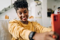 Charmante Afroamerikanerin mit lockigem Haar macht Selbstporträt auf dem Smartphone, während sie zu Hause auf dem Sofa sitzt — Stockfoto
