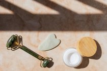 Верхний вид нефритового ролика, гуашского скребка и натурального крема для лица расположен на мраморном столе для концепции массажа лица — стоковое фото