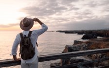Чоловік з рюкзаком і капелюхом стоїть на прогулянці, дивлячись на море — стокове фото