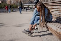 Vista lateral Afro-americana hembra en traje de mezclilla poniéndose patines en día soleado en skate park - foto de stock