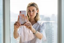 Junge Frau steht in leerem Büro mit großen Fenstern und macht Selfie mit dem Handy — Stockfoto