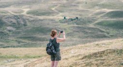 Rückansicht einer anonymen Backpackerin, die während einer Sommerreise im Sonnenlicht Hügel mit dem Handy fotografiert — Stockfoto