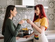 Side view of young smiling homosexual women with glasses and bottle of white wine speaking in house while looking at each other — Stock Photo