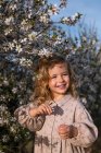 Очаровательный улыбающийся маленький ребенок в платье стоит рядом с цветущим деревом с цветами в весеннем парке и смотрит в сторону — стоковое фото