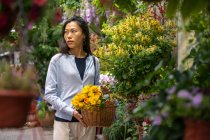 Schönes asiatisches Mädchen kauft Blumen im Blumenladen, während es einen Weidenkorb mit gelben Blumen trägt. — Stockfoto