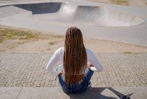 Черная женщина с заплетенной прической и в роллах сидела на рейнджерах в скейт-парке и отводила взгляд — стоковое фото