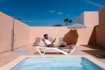Vista lateral del freelancer masculino tumbado en la tumbona junto a la piscina y navegando por Internet en el teléfono móvil durante el teletrabajo en verano en un día soleado - foto de stock