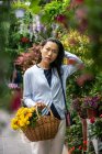 Hermosa chica asiática comprando flores en la tienda de flores mientras lleva una canasta de mimbre con flores amarillas. - foto de stock