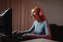 Vista laterale del gamer femminile felice in cuffie che giocano videogame mentre si siede a tavola a casa — Foto stock