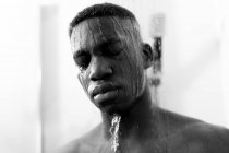Noir et blanc de jeune homme noir sans émotion prenant une douche dans la salle de bain lumineuse avec les yeux fermés et de l'eau sur le visage — Photo de stock