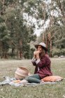 Женщина в повседневной одежде и шляпе наслаждается вкусным сэндвичем, сидя на ткани возле плетеной корзины и глядя в сторону — стоковое фото