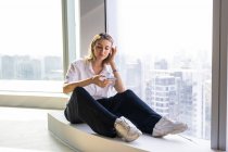 Самотня неемоційна молода жінка сидить у порожньому офісі з великим вікном, що переглядає мобільний телефон — стокове фото