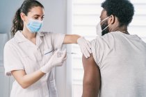 Especialista médica em uniforme de proteção, luvas de látex e máscara facial vacinando pacientes afro-americanos na clínica durante o surto de coronavírus — Fotografia de Stock