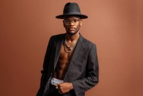 Junger männlicher unrasierter afroamerikanischer Mann mit nacktem Bauch in Jacke blickt in die Kamera auf braunem Hintergrund — Stockfoto