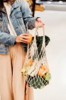 Cropped unrecognizable female standing with assorted fruits and vegetables in eco friendly mesh bag against blue wall in city — Stock Photo