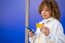 Content stylish African American female paying with plastic card during online shopping via mobile phone while standing on blue background in studio — Stock Photo
