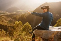 Vue latérale du voyageur masculin en lunettes VR interagissant avec la réalité virtuelle assis sur une colline en terrain montagneux au coucher du soleil — Photo de stock