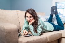 Молодая счастливая улыбающаяся женщина в бирюзовой толстовке и очках, лежащих на диване с помощью мобильного телефона — стоковое фото
