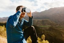 Curioso viaggiatore maschile seduto sulla collina e sperimentare la realtà virtuale in maschera in montagna nella giornata di sole — Foto stock