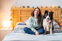 Junge Frau mit Brille sitzt auf Bett und umarmt Border Collie Hund, der in die Kamera schaut — Stockfoto
