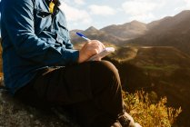Вид збоку врожаю невпізнаваний дослідник чоловічої статі сидить на пагорбі в горах і пише в блокноті під час відпустки — стокове фото