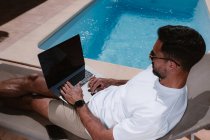 Freelancer masculino tumbado en una tumbona junto a la piscina y navegando por Internet en el portátil durante el teletrabajo en verano en un día soleado - foto de stock