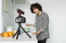 Hombre joven con camisa a rayas que habla contra la cámara fotográfica en el trípode durante el proceso de cocción en la cocina - foto de stock