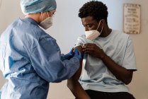 Ärztin in Schutzuniform und Latexhandschuhen impft während des Coronavirus-Ausbruchs einen afroamerikanischen Patienten in der Klinik — Stockfoto