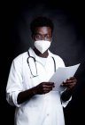 Médico afroamericano en máscara protectora y uniforme blanco tomando notas en el portapapeles mientras mira la cámara sobre fondo negro - foto de stock