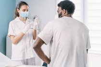 Doctora en guantes de látex y protector facial llenando jeringa de frasco con vacuna preparándose para vacunar a un paciente afroamericano irreconocible en la clínica durante el brote de coronavirus - foto de stock