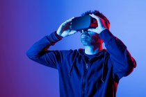 Varón barbudo irreconocible con capucha y auriculares modernos que exploran la realidad virtual con luz de neón - foto de stock