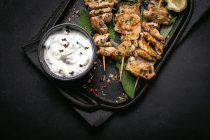 Зверху апетитне м'ясо на шампурах, подане на підносі на чорному столі з мискою соусу — стокове фото
