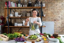 Happy female mixing tasty vegetable salad with lettuce leaves at table in loft style house — Stock Photo