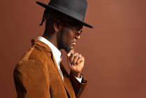 Vue latérale du jeune homme afro-américain dans des vêtements à la mode et un chapeau regardant loin sur fond brun — Photo de stock