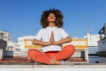 Затишна афроамериканка сидить в Падмасані з Намасте жестом і медитує закритими очима, практикуючи йогу на даху. — стокове фото