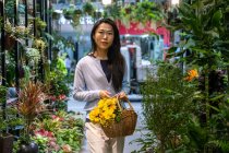 Schönes asiatisches Mädchen kauft Blumen im Blumenladen, während es einen Weidenkorb mit gelben Blumen trägt. — Stockfoto