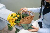 Detalhe da menina da bela asiática em um parque enquanto ela se senta e observa seu celular ao lado da cesta de vime com flores amarelas. — Fotografia de Stock