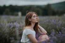 Vista lateral de hembra suave con flores en el pelo sentado en el campo de lavanda floreciente y disfrutando de la naturaleza con los ojos cerrados - foto de stock
