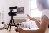 Вид збоку молодої етнічної жінки відеоблогер з блокнотом, що сидить за столом з фотоапаратом на тринозі на кухні — стокове фото