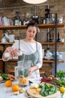 Adult female pouring vegetarian milk into bowl with chard leaves and orange slices while preparing smoothie at home — Stock Photo