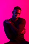 Впевнений молодий чорний чоловік в темному спортивному вбранні дивиться на камеру на неоновому рожевому фоні в темній студії — стокове фото
